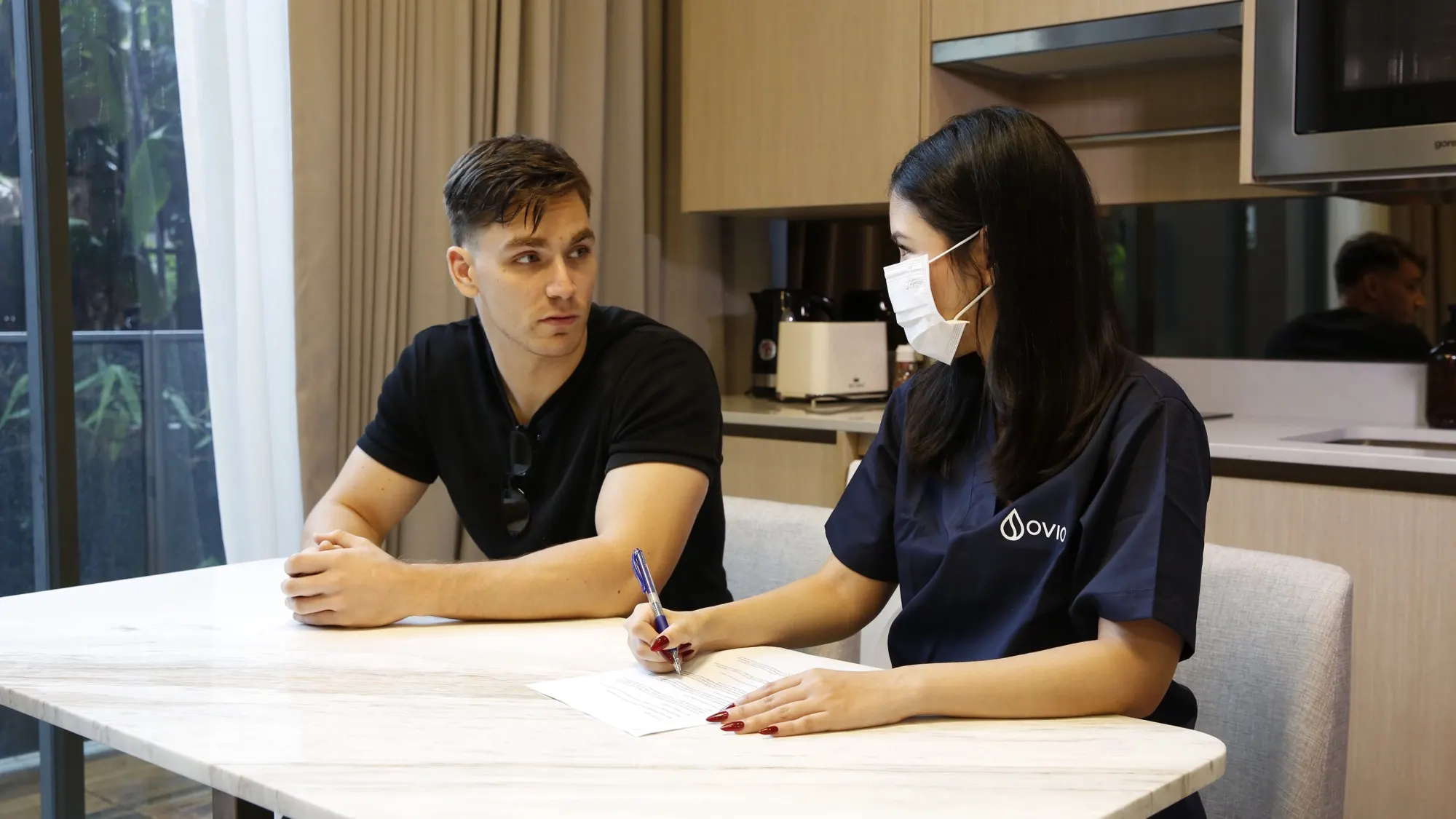 An Ovio nurse taking notes while talking with a client at his kitchen table in a Bangkok condo