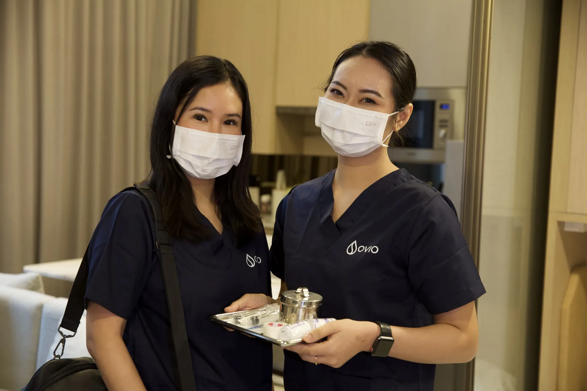 Two Ovio nurses in navy scrubs and masks smiling in a Bangkok condo, one holding a tray of sample collection supplies